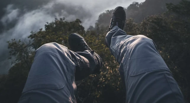 A person's legs in light pants and dark shoes dangle over a misty cliff, surrounded by lush greenery and a cloudy sky.